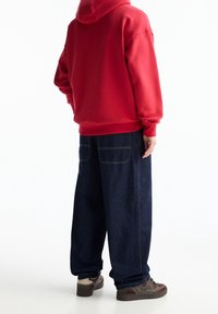 Czerwona bluza z kapturem z długimi rękawami, zestawiona z luźnymi, ciemnoniebieskimi dżinsami. Brązowe sneakersy z teksturowanym wzorem.