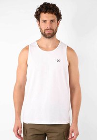 Débardeur en coton blanc avec un col rond et des emmanchures larges, arborant un petit logo géométrique noir sur la poitrine gauche.