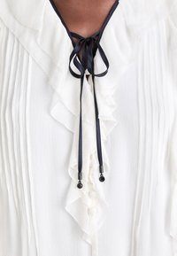 Weiße Bluse mit Rüschendetails, marineblaue Satinbänder; mit gefältelten Akzenten und dekorativen schwarzen Perlen. Knopfverschluss vorne.