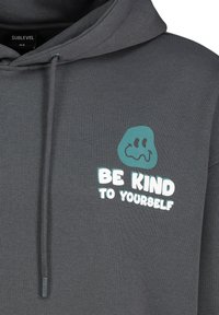 Grijze hoodie met een zachte textuur, voorzien van een teal blobgraphic en de woorden "WEES VRIENDELIJK VOOR JEZELF" in witte tekst op de voorkant.