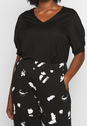 Vrouw draagt een zwarte top met V-hals en pofmouwen tot aan de ellebogen en zwarte hoge taille broek met wit abstract penseelstrekenpatroon
