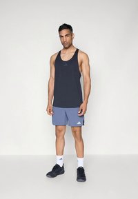 Débardeur de sport en tissu bleu foncé avec un col rond, associé à un short bleu clair avec un logo Adidas ; porté avec des chaussures de sport noires.