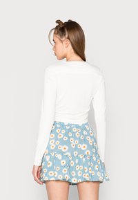 Top branco de mangas compridas combinado com uma saia mini azul com padrão de margaridas e bainha em rendado, exibindo uma textura leve de algodão.