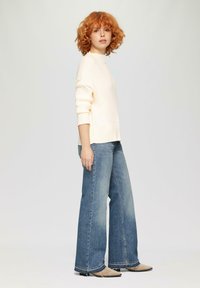 Helles Pfirsichfarbnes Rippenpullover mit umgeschlagenen Ärmeln, kombiniert mit weit geschnittenen blauen Jeans. Das Model trägt beigefarbene spitz zulaufende Schuhe.