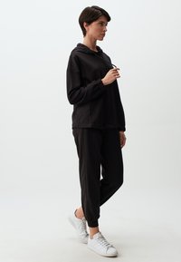 Zwarte hoodie met trekkoord en elastische manchetten, gecombineerd met bijpassende joggingbroek. Model draagt witte sneakers. Glad materiaal, relaxed fit.