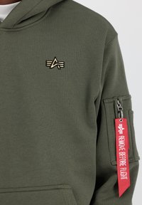 Felpa con cappuccio color khaki in cotone, con un logo dorato sul petto, tasca con zip sulla manica e un'etichetta rossa "RIMUOVERE PRIMA DEL VOLO".
