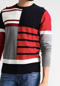 Pull en maille multicolore avec des rayures audacieuses et des blocs de couleur en marine, rouge, gris et blanc, avec des poignets et un ourlet côtelés.