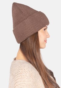 Braune gestrickte Mütze mit geripptem Muster, spitzem oberen Ende und umgeschlagenem Rand; getragen von einer Person mit langen Haaren vor einem einfarbigen Hintergrund.