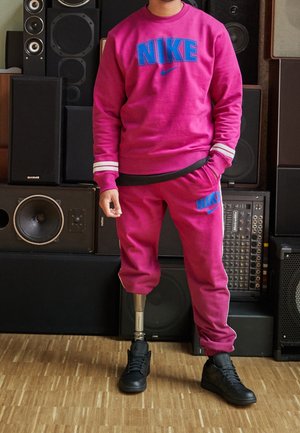 Osoba ubrana w jaskraworóżowy dres Nike z czarnymi butami, stojąca z protezą nogi przed ułożonymi na stosie głośnikami audio.