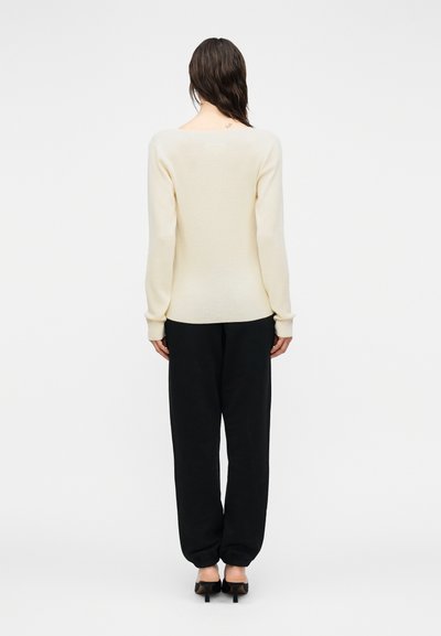 Πλεκτό πουλόβερ σε κρεμ χρώμα με φαρδιά λαιμόκοψη, συνδυασμένο με φαρδιά μαύρη φόρμα και μαύρα παπούτσια με τακούνι.