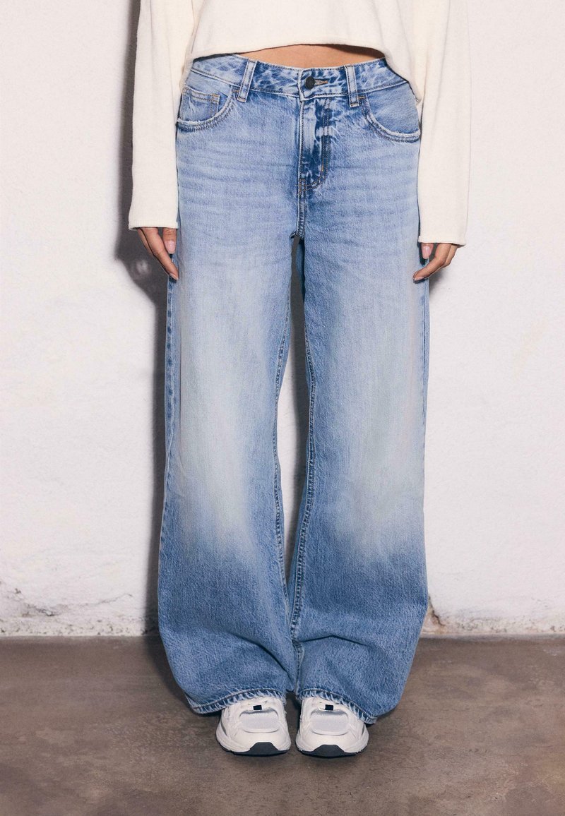 Personne portant un jean large bleu clair et des baskets blanches, debout contre un mur uni avec les mains relâchées sur les côtés.