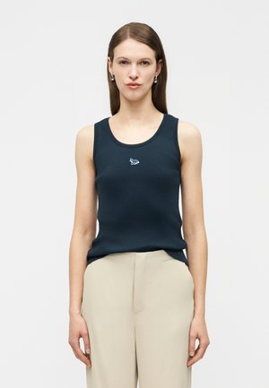 Femme portant un débardeur bleu marine avec un petit logo animalier et un pantalon beige taille haute, debout devant un fond uni.