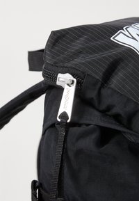 Svart ryggsekk med glidelås, strukturert overflate med fine striper og en sølvfarget glidelåsgrep merket "Jansport."