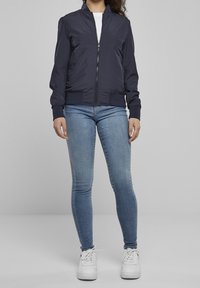 Veste bomber bleu marine avec fermeture éclair, poignets et ceinture côtelés, associée à un jean skinny bleu clair et des baskets blanches.