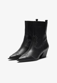 Botas de cuero negro hasta el tobillo con punteros afilados, detalles de tachuelas y un tacón bajo tipo bloque. Presentan un diseño de costura decorativa y una cremallera lateral.