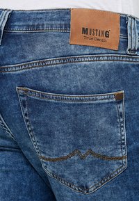 Jeans dżinsowe z przetarciami w niebieskim odcieniu, z skórzaną metką oznaczoną "Mustang True Denim" i kontrastującym żółtym szwem na tylnej kieszonce.