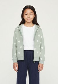 Felpa verde chiaro con zip e motivi a cuori bianchi, dotata di cappuccio, tasche frontali e polsini a coste, abbinata a pantaloni plissettati blu navy.