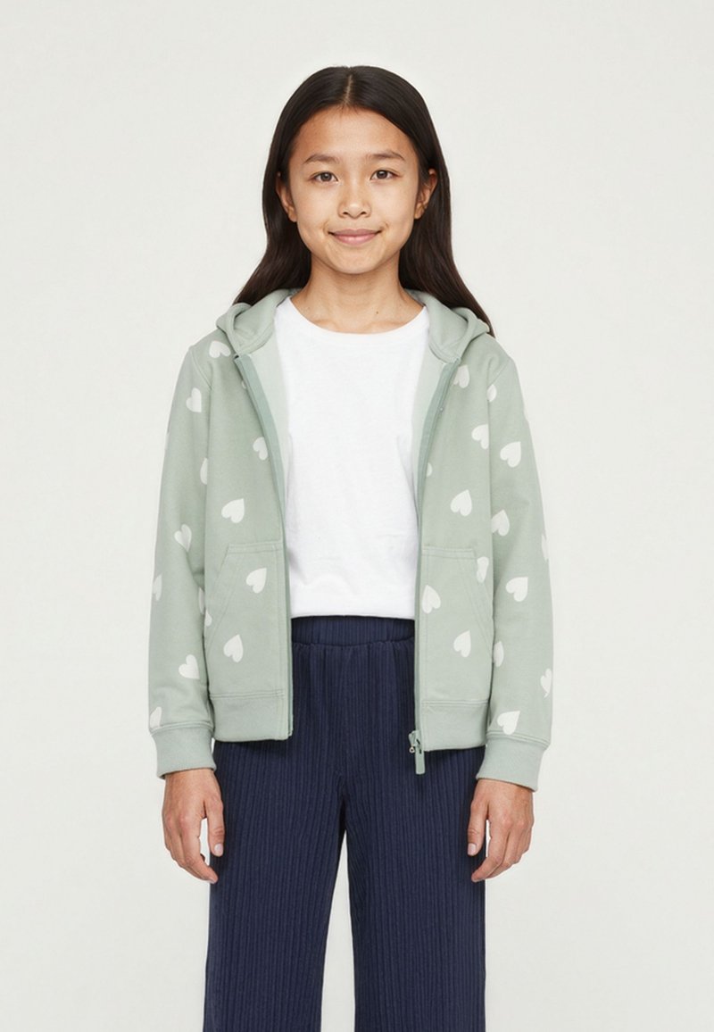 Felpa verde chiaro con zip e motivi a cuori bianchi, dotata di cappuccio, tasche frontali e polsini a coste, abbinata a pantaloni plissettati blu navy.