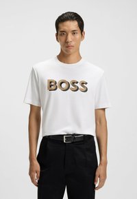 Młody mężczyzna w białej koszulce z napisem "BOSS" w beżowym i czarnym kolorze, z czarnym paskiem i czarnymi spodniami na jednolitym białym tle.