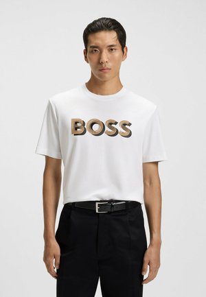 Young man wearing a white T-shirt with "BOSS" text in beige and black, black belt, and black pants against a plain white background.