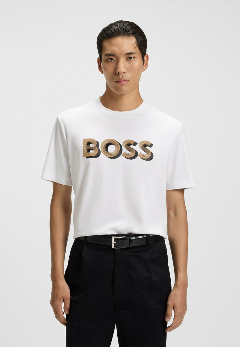 Młody mężczyzna w białej koszulce z napisem "BOSS" w beżowym i czarnym kolorze, z czarnym paskiem i czarnymi spodniami na jednolitym białym tle.