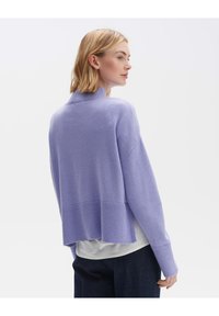 Maglione lavorato a maglia di colore viola chiaro con colletto alto, spalle scese, orlo a costine e spacchi laterali, indossato sopra una camicia bianca.