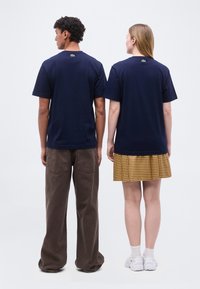 Άνδρας και γυναίκα στέκονται πλάτη φορώντας μπλε σκούρες κοντομάνικες μπλούζες, καφέ παντελόνια και κίτρινη καρό φούστα σε λευκό φόντο.