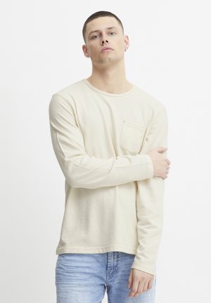 Long-sleeve beige shirt made of textured fabric with a rounded neckline and chest pocket; paired with light blue jeans.