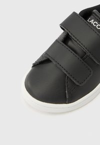 Μαύρο συνθετικό παπούτσι με στρογγυλεμένη μύτη, διαθέτει δύο ιμάντες με βέλκρο και λευκή λαβή από καουτσούκ. Ομαλή υφή με ραφές ως διακοσμητικά στοιχεία.