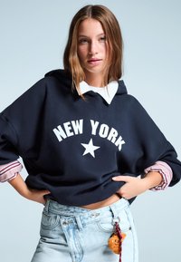 Námořnická mikina s bílým textem "NEW YORK" a hvězdičkovou grafikou, navrstvená přes bílou košili s límečkem. Světle modré denimové šortky s přívěškem na klíče.