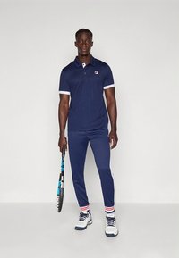 Mornarsko modra polo majica z belimi poudarki, usklajene mornarsko modre športne hlače, beli športni copati, drži teniški lopar. Oblikovanje priložnostne športne opreme.