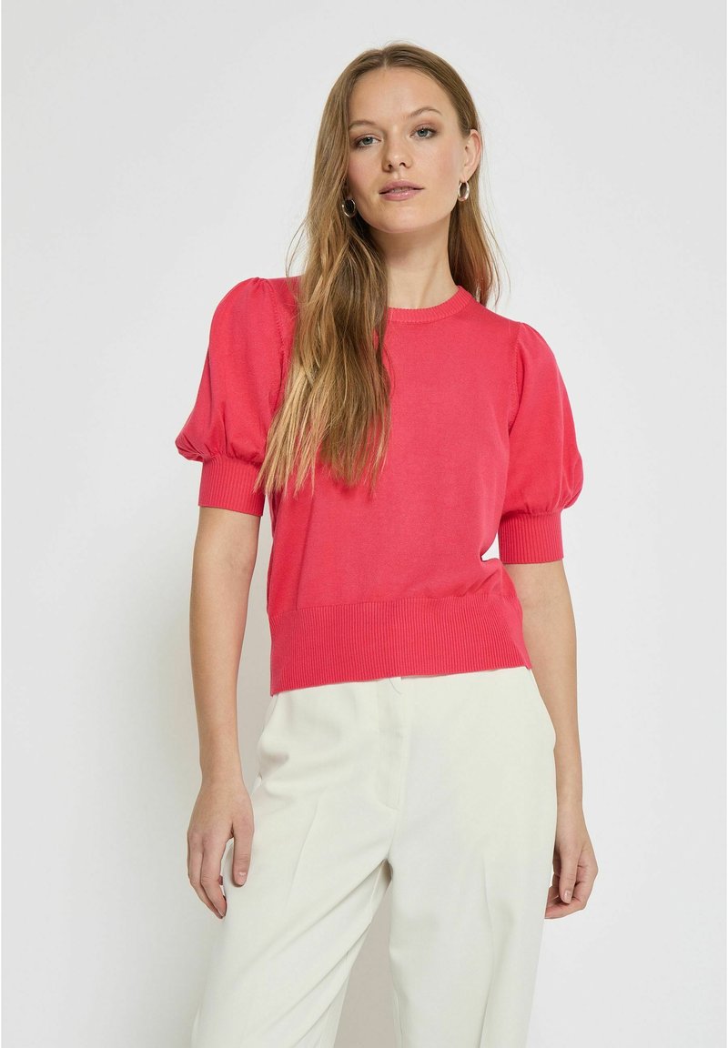 Pull en tricot corail avec des manches bouffantes, un col côtelé et un ourlet côtelé. Associé à un pantalon clair et ample. Texture lisse.