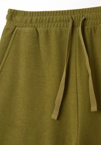 Olivgrüne Jogginghose mit strukturierter Oberfläche, ausgestattet mit einem elastischen Bund und einer Zugkordel zur Anpassung. Zwei seitliche Taschen sind sichtbar.