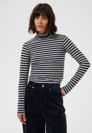 Femme aux cheveux foncés mi-longs portant un col roulé rayé noir et gris et un pantalon taille haute foncé, posant sur un fond uni.