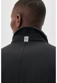 Giacca nera con colletto alto, tessuto morbido e marchio visibile sul retro. Il design include linee pulite e accenti minimi.