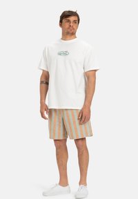 T-shirt bianca in cotone con una piccola grafica colorata a tema marino. Abbinata a pantaloni corti a righe tan e teal e scarpe da ginnastica bianche.