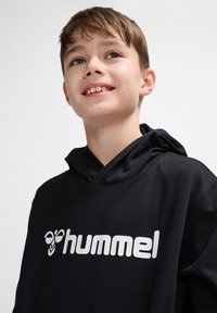 Schwarzer Hoodie aus weichem Stoff mit einem großen weißen „hummel“-Logo. Die Kapuze hängt locker mit einem weiten Ausschnitt und subtiler Textur.