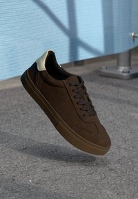 Brown suede low-top sneakers with black laces, featuring a white heel cap and textured stitching details. Rubber sole.
