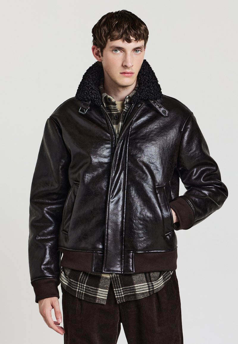 Dunkelbraune Lederbomberjacke mit einem strukturierten schwarzen Pelzkragen, Reißverschluss und gerippten Bündchen. Getragen über einem karierten Hemd.