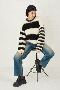 Črno-bela prugasta puhasta pulover, prevelikega kroja. Par svetlih ravnih jeansov in masivnih črnih superg. Sedi na kovinskem stolu.