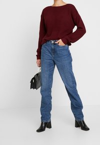 Ribbeliger burgunderfarbener Pullover mit langen Ärmeln, kombiniert mit hellblauen geraden Jeans. Das Model hält eine kleine schwarze Handtasche und trägt schwarze Absatzstiefel.