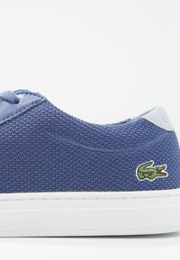 Baskets en tissu bleu marine avec un dessus en maille texturée, un accent lisse au niveau du talon et une semelle en caoutchouc blanc ; présente un logo de crocodile brodé en vert.
