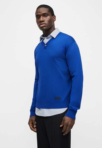 Άνδρας που φοράει φωτεινό μπλε πουλόβερ με μακριά μανίκια πάνω από ένα ανοιχτόχρωμο ριγέ πουκάμισο με γιακά και μαύρο παντελόνι, στέκεται μπροστά σε απλό ανοιχτό φόντο.