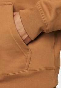 Felpa marrone con una superficie texture, dotata di tasca laterale e polsini a coste. Primo piano di una mano appoggiata nella tasca.