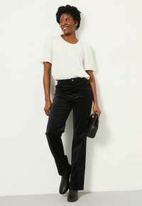 Weißes Kurzarmhemd mit dezenten Verzierungen, kombiniert mit schwarzen Cordhosen im Schlag und schwarzen Ankle Boots. Hält eine schwarze Tasche.