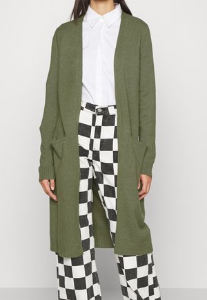 Kvinde iført en lang olivengrøn cardigan over en hvid skjorte med knapper og sort-hvide ternede bukser, stående mod en ensfarvet baggrund.