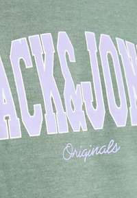 Tissu vert avec un grand texte blanc en contour "JACK & JONES" et un texte cursif plus petit "Originals" en dessous.