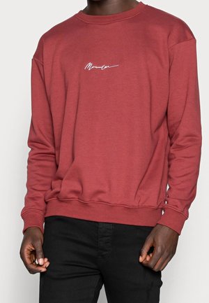 Homme portant un sweat-shirt à manches longues couleur rouille avec un texte blanc sur la poitrine et un pantalon noir sur un fond uni.