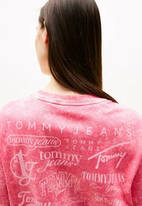 Różowy sweter z miękkiego materiału, z różnymi białymi logo i wzorami, pokazujący zużyty, vintage'owy styl. Okrągły dekolt, luźny krój.