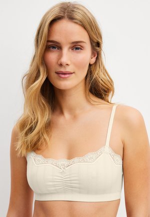 Blonde Frau mit welligem Haar, die ein cremefarbenes Spitzen-Bralette trägt, blickt mit neutralem Gesichtsausdruck nach vorne vor einem schlichten Hintergrund.